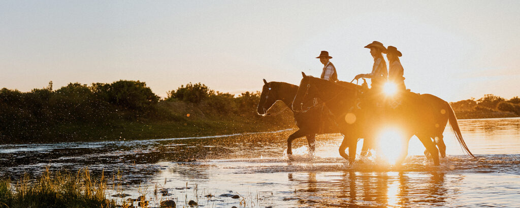 Three women in cowboy hats ride horses through water.